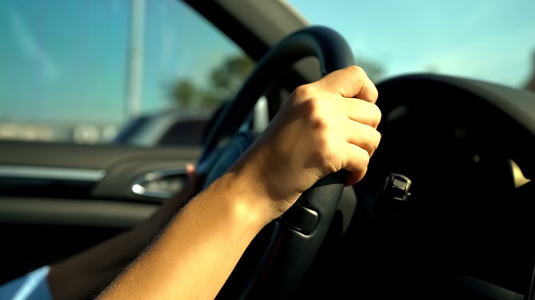 Teenager’s hands gripping a steering wheel, with bright sunlight streaming into the car and a clear view of the side window and dashboard.