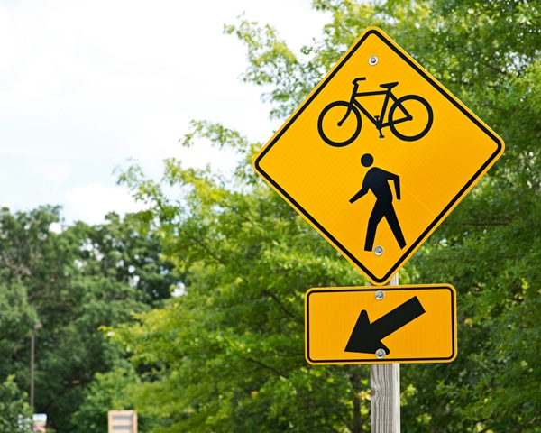 Yellow caution sign showing symbols for a pedestrian and a bicycle above a directional arrow, indicating a shared path or crossing area to the left for walkers and cyclists.