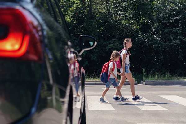 Two children with backpacks crossing a crosswalk in front of a stopped car on a sunny day.