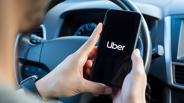 A person in the driver's seat of a car holds a smartphone displaying the Uber logo.
