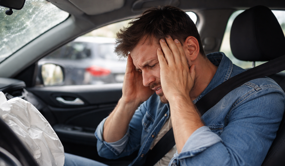 A man in a denim shirt sits in the driver's seat of a car with a deployed airbag, grimacing in pain and holding both hands to his head.