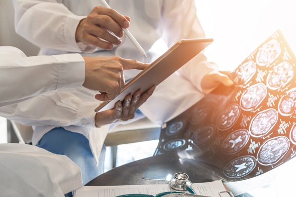 Two medical professionals in white coats reviewing a digital tablet and a large sheet of brain MRI scans, with a stethoscope and medical paperwork visible on a table in the foreground.