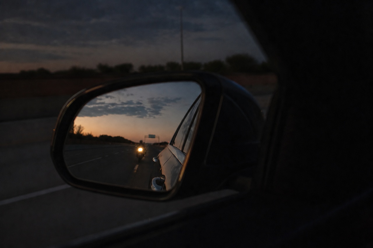 A view from inside a vehicle looking through the side-view mirror, which reveals a motorcyclist riding closely behind in the blind spot, illustrating the critical driver visibility issues that contribute to Texas motorcycle crashes.