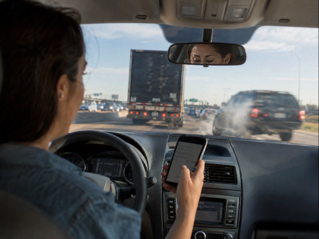 A female driver is seen from the backseat holding a smartphone in her right hand while driving on a busy highway, with her reflection in the rearview mirror showing her looking down at the device instead of the road ahead, where a large semi-truck and other vehicles are in close proximity.